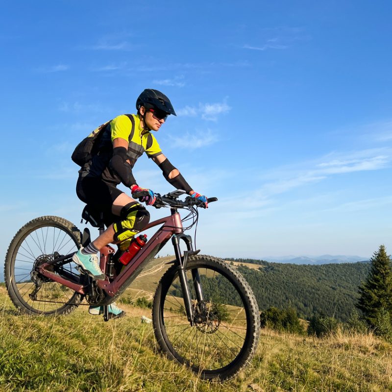 Cyclist man riding electric mountain bike outdoors. Male tourist biking along grassy trail in the mountains, wearing helmet and backpack. Concept of sport, active leisure and nature.
